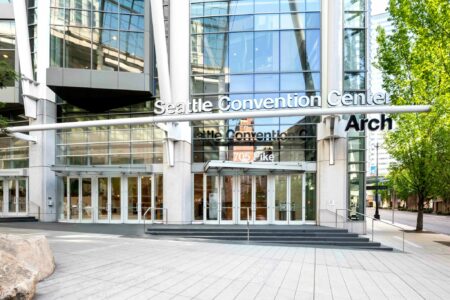 Entrance of the Seattle Convention Center, featuring large glass windows and the name displayed above the door. The area is well-lit with a modern design, and there are trees visible on the right side.