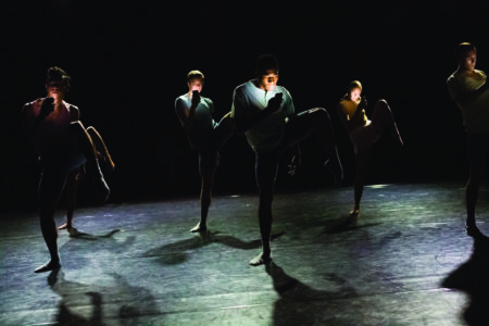 A group of dancers perform on a dimly lit stage, each lifting one knee in unison. Their faces are partially shadowed, and they wear dark, fitted clothing. The spotlight creates dramatic contrasts, highlighting their synchronized movements.