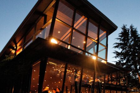 A modern, multi-story building with large windows is illuminated warmly from within. The evening sky is a deep blue, and tall trees are partially visible around the building.