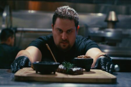 A chef wearing black gloves leans forward over a kitchen counter, preparing to serve food on a wooden board. He is focused on his work, with bowls and garnished meat visible in the foreground. The kitchen background is slightly blurred.