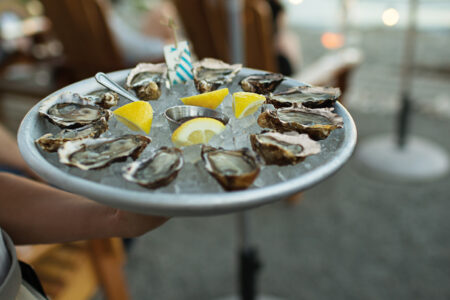 A person holds a round platter of fresh oysters on ice, garnished with lemon wedges and a small container of sauce. The background shows outdoor seating by a waterfront with blurred boats and lights.