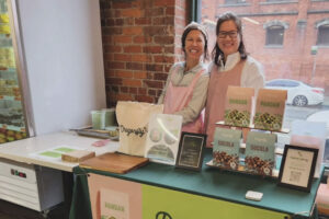 Two smiling women wearing pink aprons stand behind a table displaying pandan-flavored treats, a tote bag, framed certificates, and product boxes at an indoor market with a brick wall and large window.