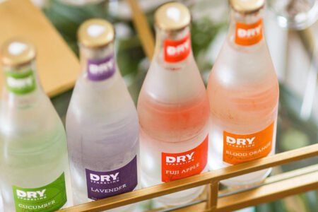 Four frosted glass bottles of DRY sparkling beverages sit in a row. Flavors are labeled as cucumber, lavender, Fuji apple, and blood orange, each with a matching colored label and cap.