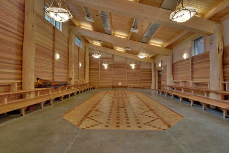Spacious wooden room with a detailed patterned wooden floor, high ceiling, and unique round chandeliers. Wooden benches line the walls, and there are decorative items, including a canoe, on the left side. Natural light filters through windows.