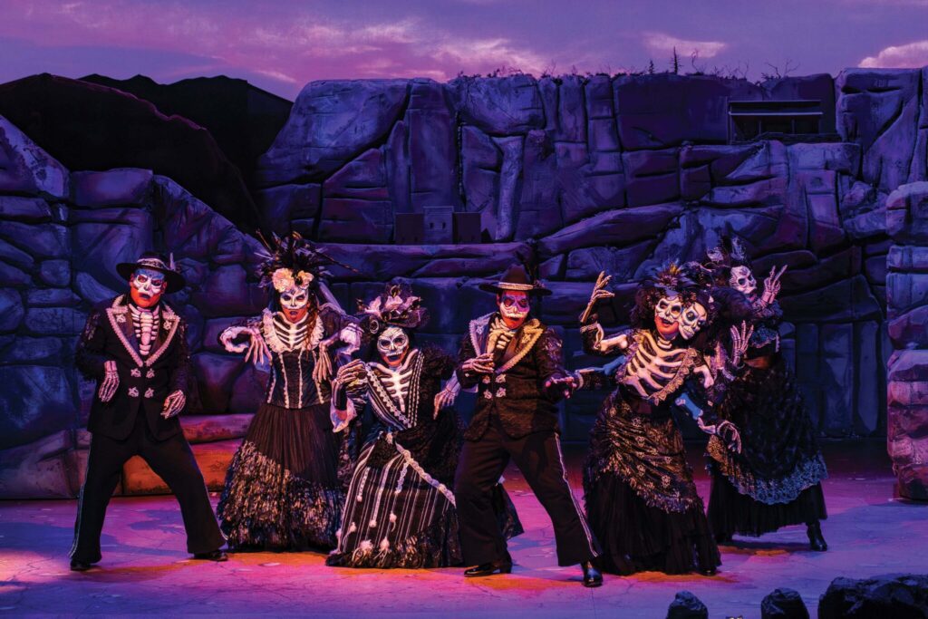 A group of performers dressed as skeletons with elaborate costumes and face paint stand on stage against a rocky backdrop. They are posing dynamically, with purple lighting casting an eerie glow.