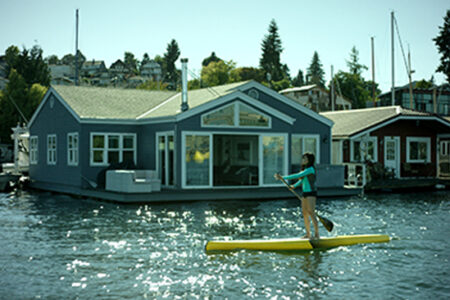 A person in a green shirt is paddleboarding on a yellow board in front of a blue houseboat on a sunny day. The water is sparkling, and trees and other houses are visible in the background.