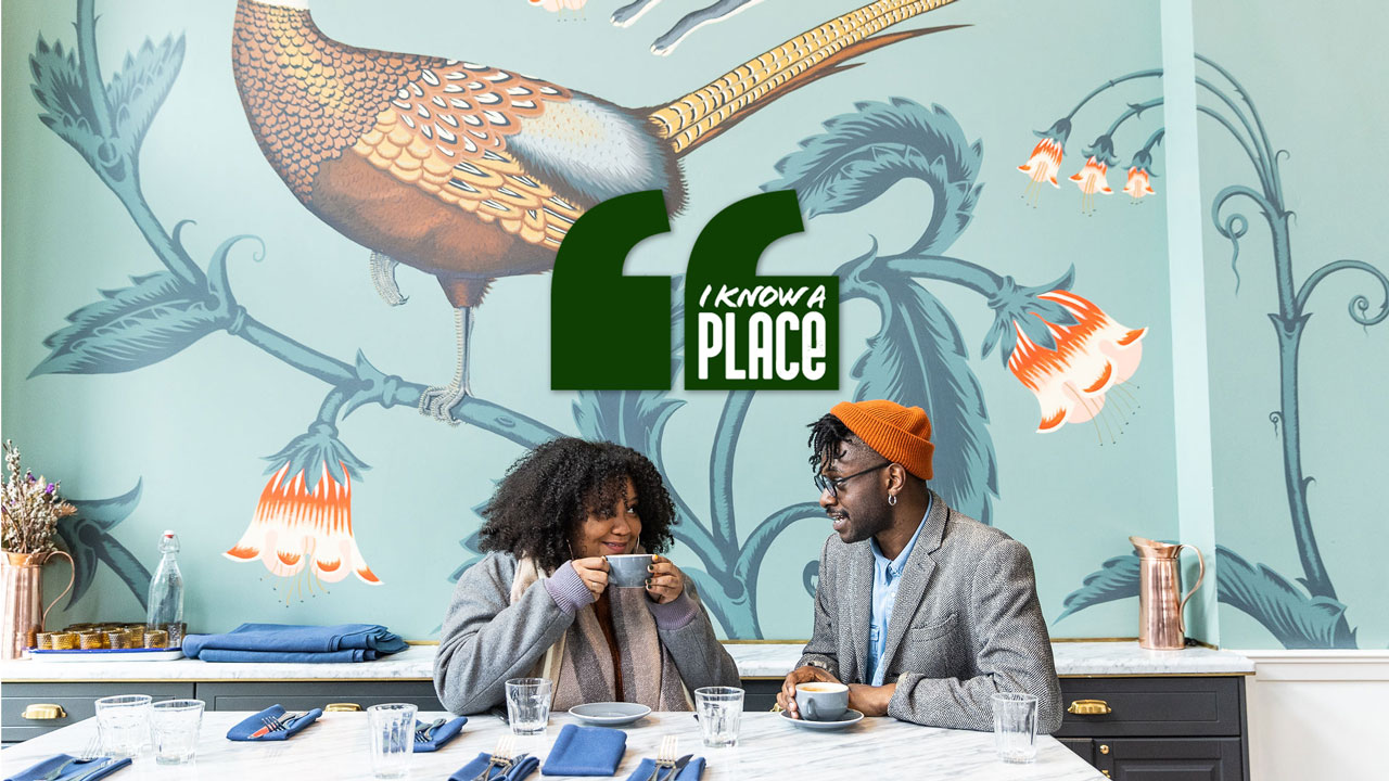 A couple enjoying coffee at a marble table in a cafe. The wall behind them is decorated with a large bird and floral design. A green sign with the text "I know a place" is displayed above them.
