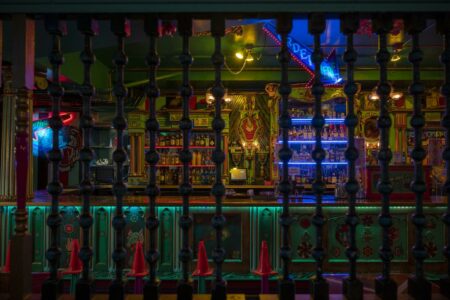 A vibrant, colorful bar interior with eclectic decor, featuring bright neon lights and unusual ornaments. The foreground has a railing of turned wooden balusters, partially obscuring the view of the illuminated bar shelves and counter.