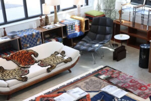 A modern living room with large windows, a white sofa with two tiger-patterned throws, a black leather lounge chair, books, colorful blankets, and various wooden shelves and tables with lamps and decor items.