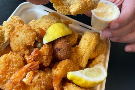 A person holds a crispy, fried piece of seafood dipped in sauce above a container filled with fried shrimp, fish, hush puppies, crinkle-cut fries, and lemon wedges. A small cup of sauce is in the background.