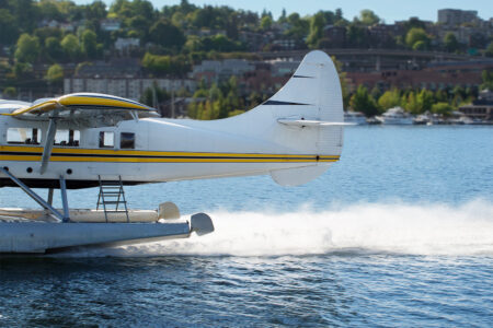 A white seaplane with yellow stripes taxis on a calm blue lake, creating a trail of water spray. A lush green shoreline and a hilly urban landscape are visible in the background under a clear blue sky.