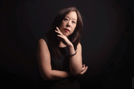 A woman with long dark hair and a sleeveless black top poses against a dark background. She rests her chin on one hand, looking thoughtfully at the camera.