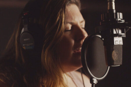Musician Mary Lambert singing into a microphone with her eyes closed against a dark background.