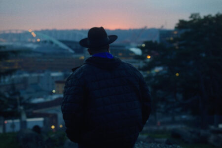 A person wearing a hat and puffy jacket stands on a hill, facing away and overlooking a cityscape at dusk. The background features blurred city lights and a bridge under a colorful evening sky.