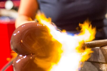 A glassblower shapes a molten glass heart with a torch. The glowing piece is being heated, showcasing vibrant orange flames. The artisan's hands are visible in the background, focusing on the intricate craft.