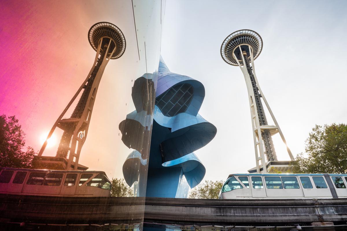 The Space Needle and a monorail in Seattle are reflected in a colorful, shiny surface next to a curved, blue modern building under a bright sky.