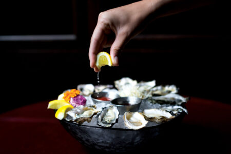 A hand squeezes a lemon wedge over a plate of oysters on ice. The dish is garnished with other lemon wedges and a small container of sauce. The scene is set against a dark background, highlighting the freshness of the seafood.