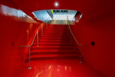 A vibrant red staircase leads upward, framed by smooth walls and metal railings. The ceiling opening reveals a glimpse of a brightly lit area with geometric patterns.