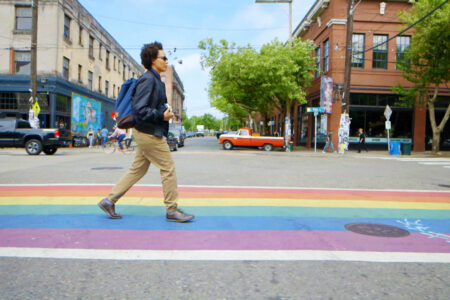 A person with curly hair and a backpack walks across a rainbow-colored crosswalk in an urban area. Surrounding buildings are lined with trees, and several parked vehicles are visible. The sky is clear with overhead cables crossing the street.