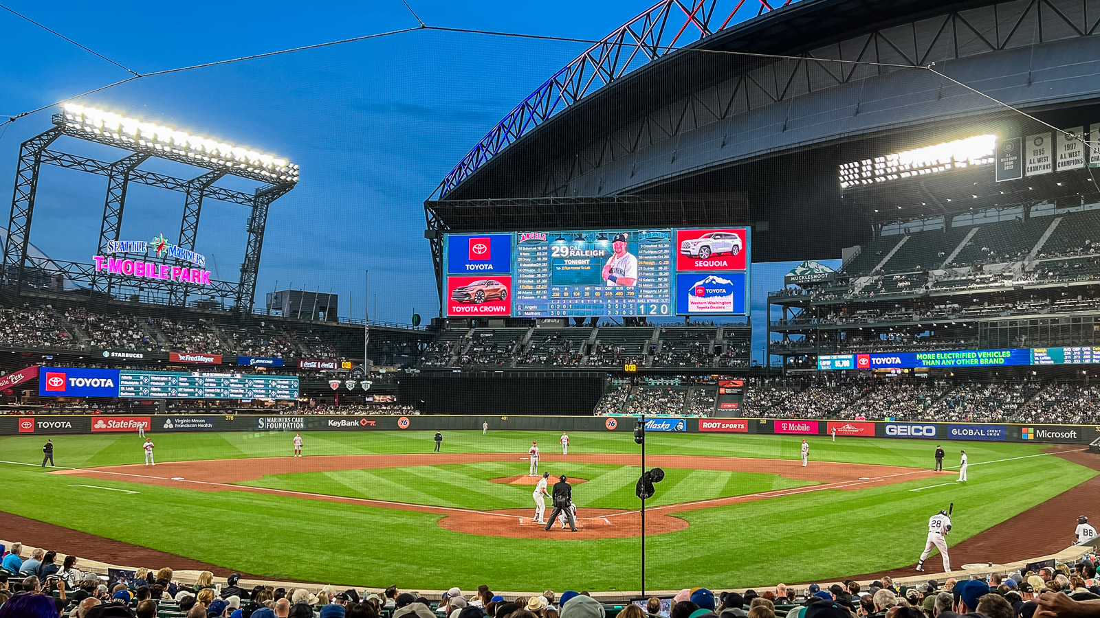 A photo taken from right behind home plate at T-Mobile Park as a Seattle Mariner is up to bat. The opposing team is in the field and a large TV screen depicts who is up to bat plus advertising on the left and right in red and blue. The lights of the stadium on the left glow against the dusky blue sky.