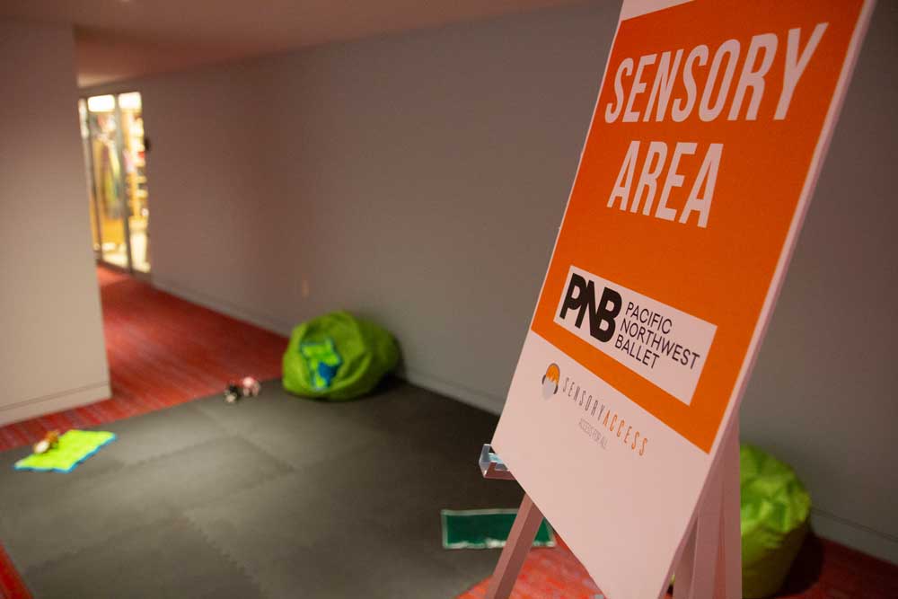 A sign reading "Sensory Area" with Pacific Northwest Ballet branding is displayed on an easel in a hallway. The area includes colorful bean bags and mats on a red carpet with soft lighting in the background.