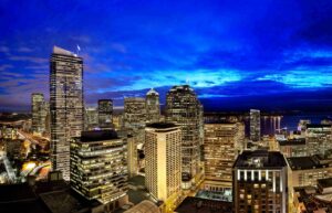 A nighttime cityscape featuring illuminated skyscrapers under a vibrant blue sky. The tall buildings are brightly lit, contrasting against the dark evening. In the background, faint hints of water and distant lights are visible.