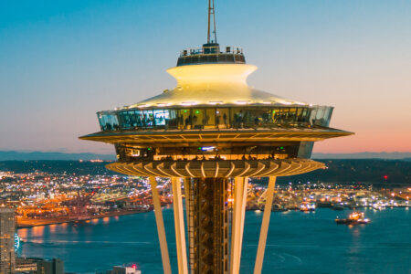 View of the Space Needle at dusk with city lights of Seattle and water in the background. The sky is clear with soft evening hues, highlighting the iconic structure's unique architecture.