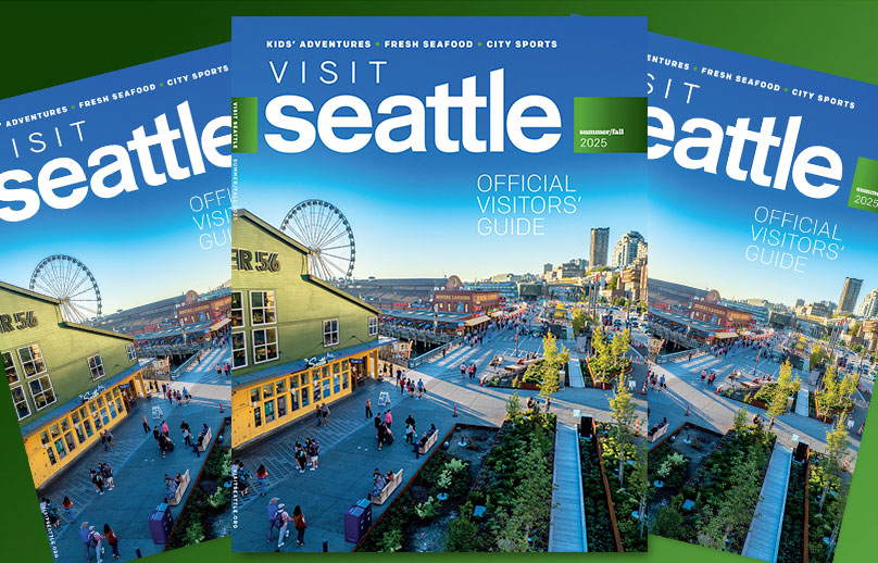 Pick up the "Visit Seattle Official Visitors’ Guide 2025" magazine, featuring a sunny view of Seattle’s waterfront with the Great Wheel, buildings, and people walking outside.