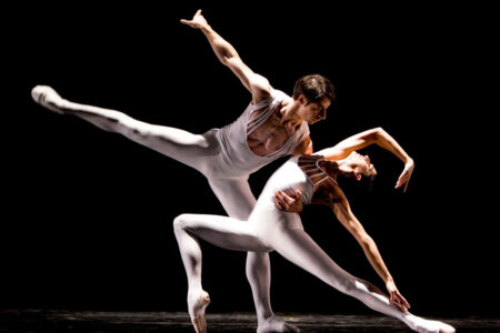In a mesmerizing display, two ballet dancers in white costumes captivate the audience on stage, silhouetted against a black backdrop. As part of the must-see things to do in Seattle, the male dancer lifts his partner effortlessly, her arm extended gracefully in an elegant pose.