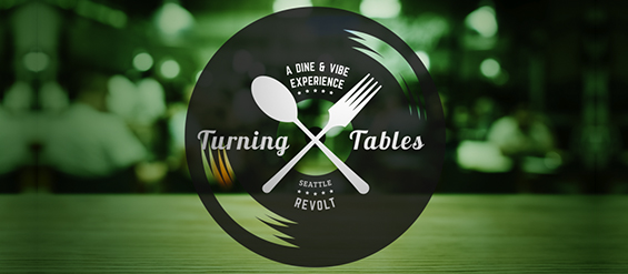 A logo with a black circular background featuring a spoon and fork crossed over each other. The text reads "A Dine & Vibe Experience" above "Turning Tables" and "Seattle Revolt" below. The background is a blurred setting with green tones.