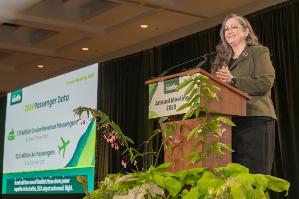 A woman with long gray hair speaks at a podium labeled "Seattle Annual Meeting 2025." A screen behind her displays "2024 Passenger Data" with cruise and air passenger stats. Green plants decorate the stage.