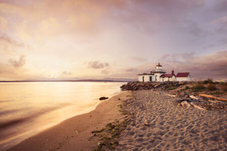 A picturesque coastal scene at sunset featuring a lighthouse on a sandy beach. The sky is filled with soft, colorful clouds, reflecting on the calm water. Driftwood and grasses line the shore leading to the lighthouse.