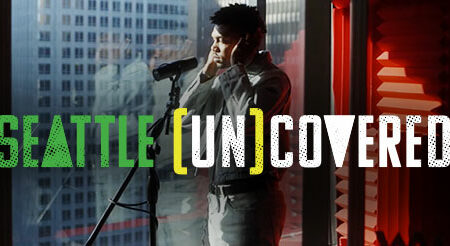 A person stands in front of a microphone in a modern studio with city views. The text "Seattle [Un]Covered" is overlaid on the image.