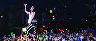 A person in a white tank top and striped pants dances on someone's shoulders at a lively nighttime outdoor concert, surrounded by a joyful crowd tossing colorful beach balls.