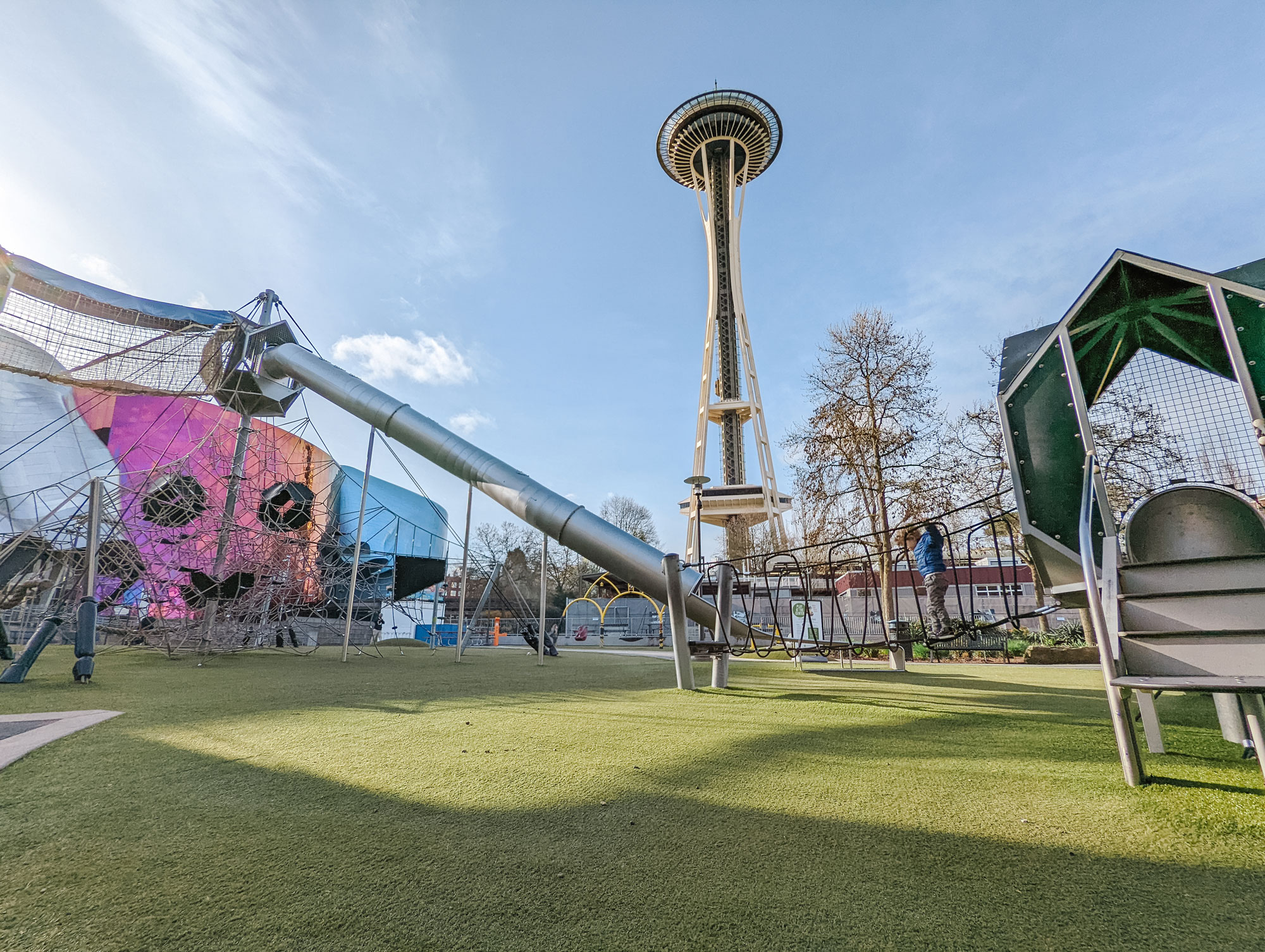 A tall and long metal slide and climbing structure are shown on the left and middle of the image. A kid walks across a black net climbing bridge that connects to a silver metal play structure with a green angular roof on the right and middle of the image. Green turf covers the floor of the playground. The colorful MoPOP museum is in the background on the left. The Space Needle rises above the playground in the middle.