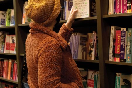 A person in a brown furry jacket and yellow beanie browses books in a bookstore. They are reaching for a book on a shelf filled with various titles.