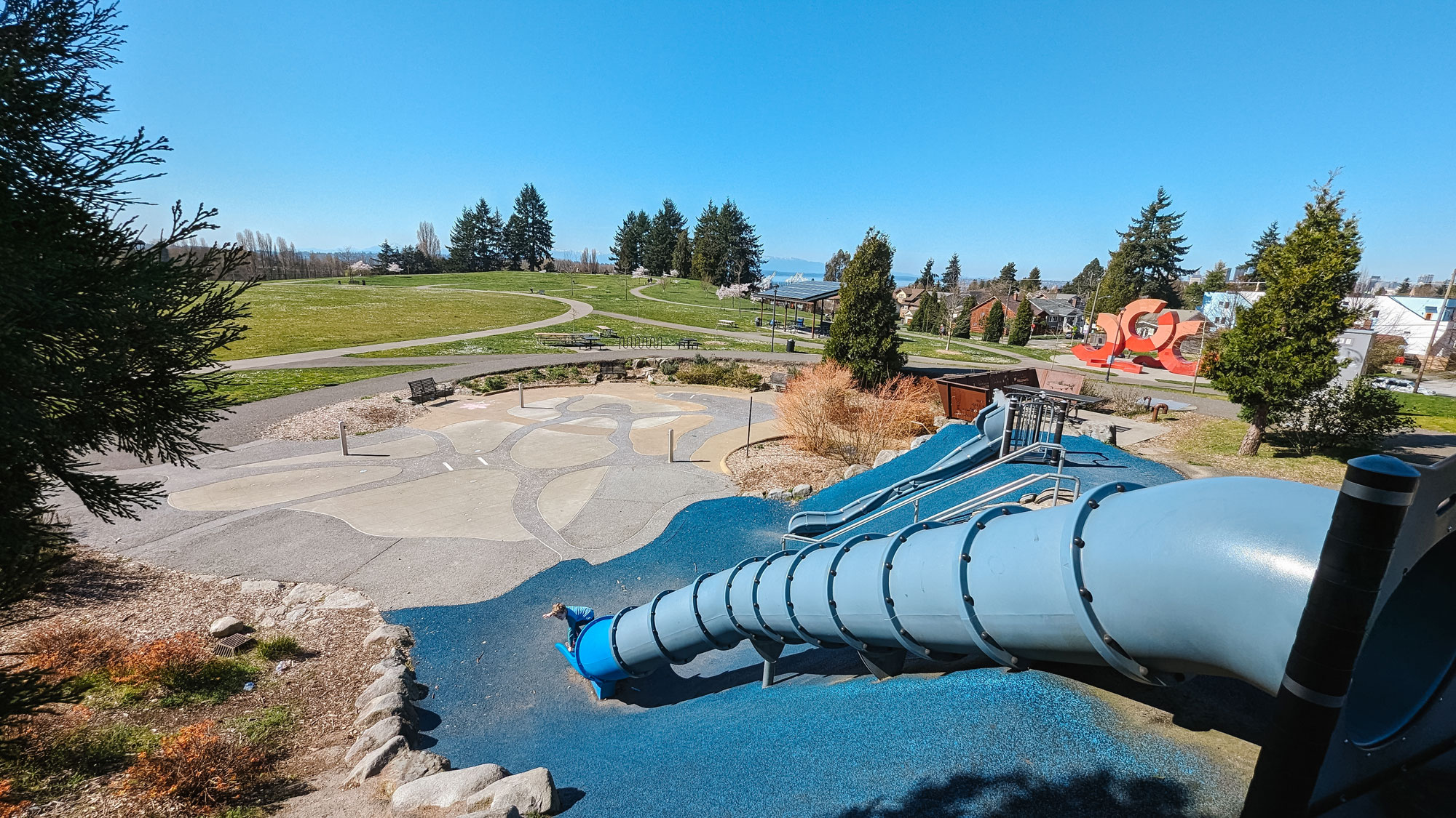 A long blue tube slide extends from the bottom right of the image. The slide ends on blue rubber playground floor. A white child wearing a blue shirt is at the bottom of the slide. A second, smaller blue slide is to the right of the primary slide. A green field with a walking path, trees, and houses are visible in the background. A large sculpture with orange rings is in the middle right of the photo.