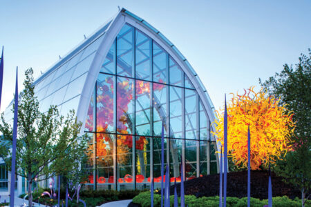 A modern glass building with an arched roof houses colorful glass sculptures in the heart of the Queen Anne neighborhood. A striking orange and yellow sculpture resembling flames stands outside, surrounded by lush green trees and shrubs.