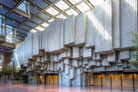 A spacious, sunlit hall features a dramatic, geometric concrete wall with angular protrusions. The hall has large ceiling panels allowing natural light to stream in, creating patterns on the floor. Doors labeled Exhibit Hall 4A are visible at the bottom.