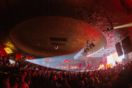 A vibrant concert venue packed with a large crowd, illuminated by colorful stage lights. The audience stands beneath a textured, dome-shaped ceiling, facing a bright stage. Energetic atmosphere filled with excitement.